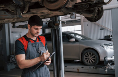 Under hanged vehicle. Man at the workshop in uniform using notepad for his job for fixing broken car.